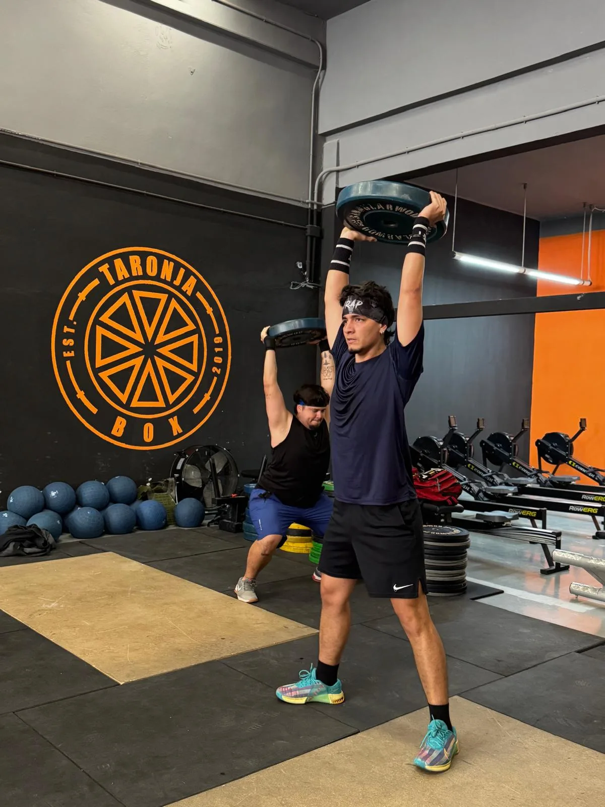 Atleta realizando overhead press con disco junto al logo de Taronja Box en Valencia