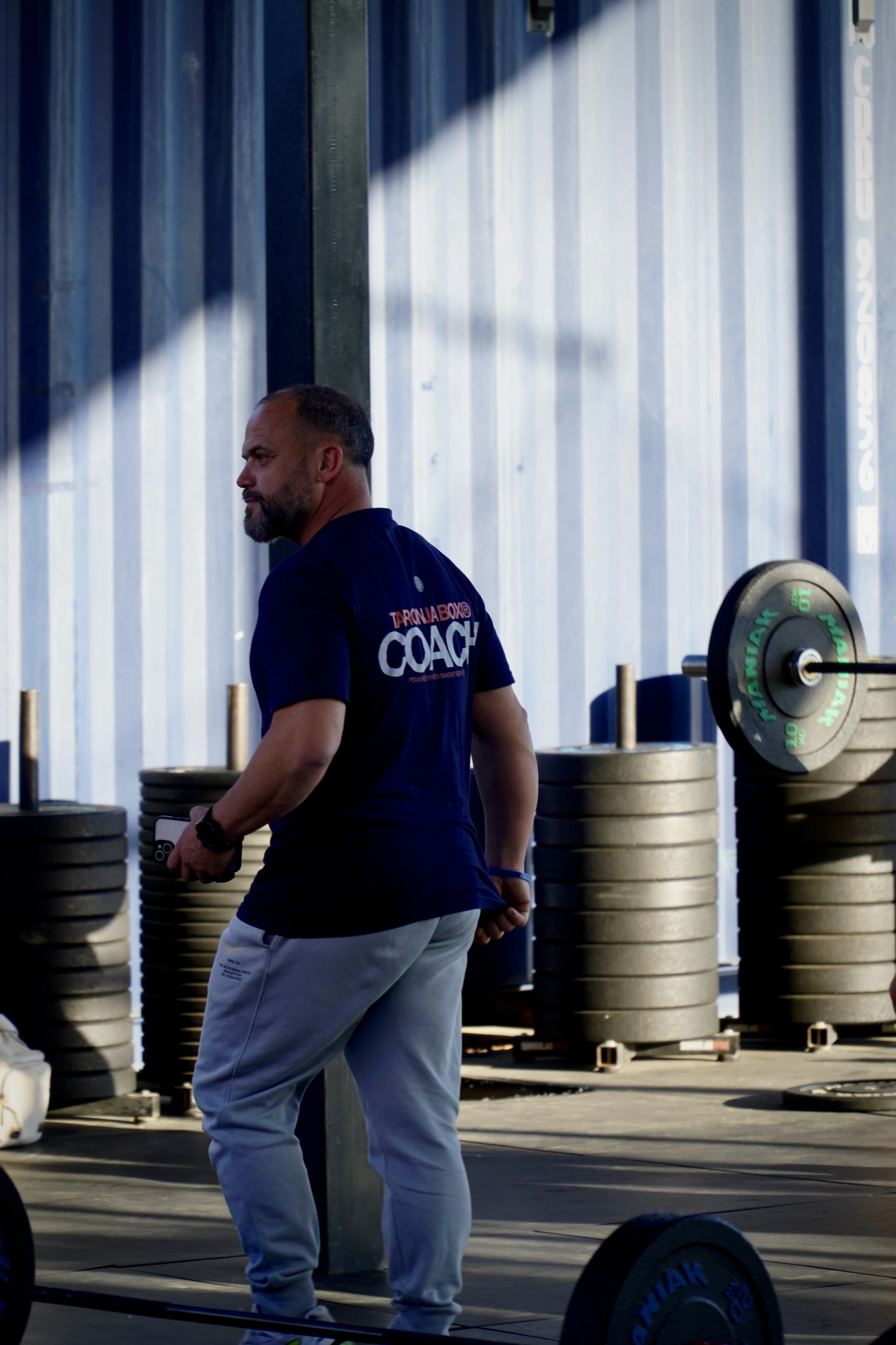 Coach con camiseta del logo junto a discos bumper plate de halterofilia en Taronja Box
